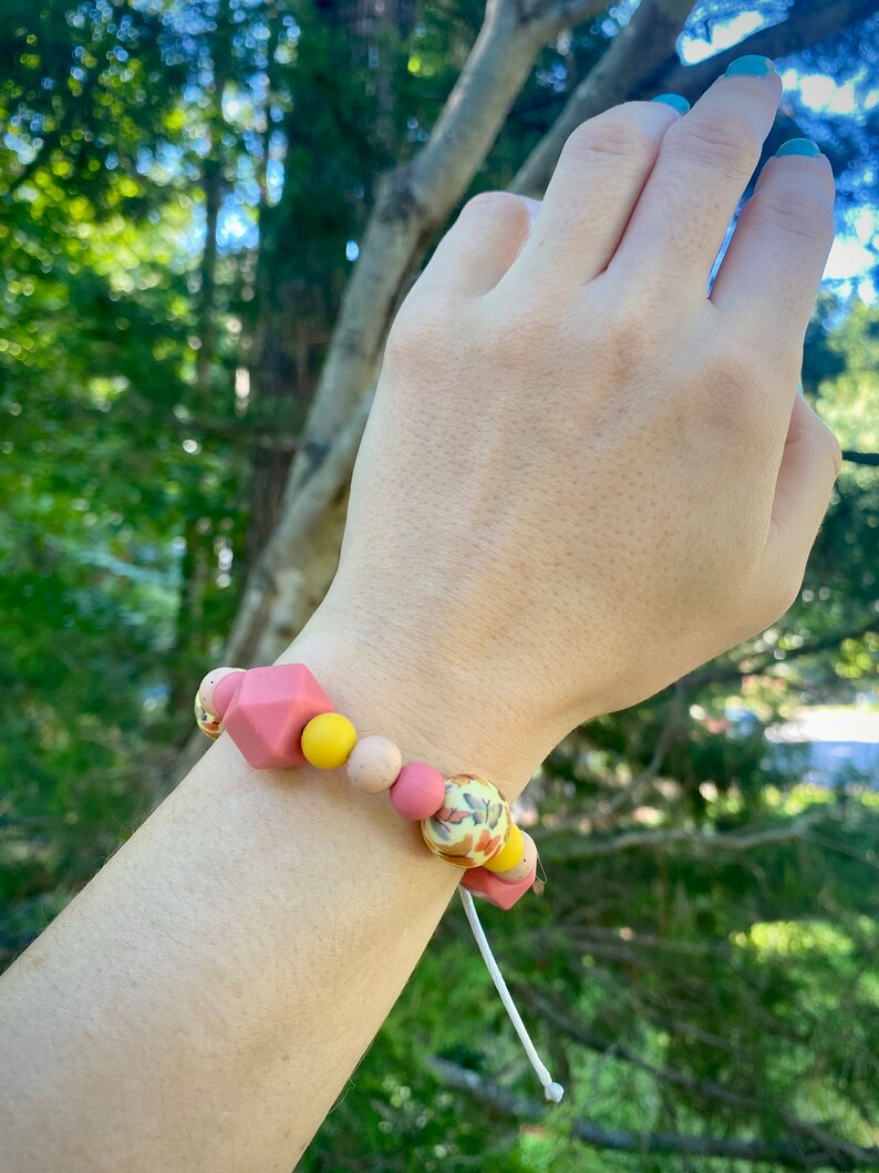 May include: A pink and yellow silicone bracelet with a floral pattern bead. The bracelet is on a person's wrist.