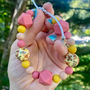 May include: A pink and yellow silicone teething bracelet with a variety of textures and patterns. The bracelet has a knotted cord and is strung with beads that include a geometric shape, a patterned bead, and a solid colour bead.