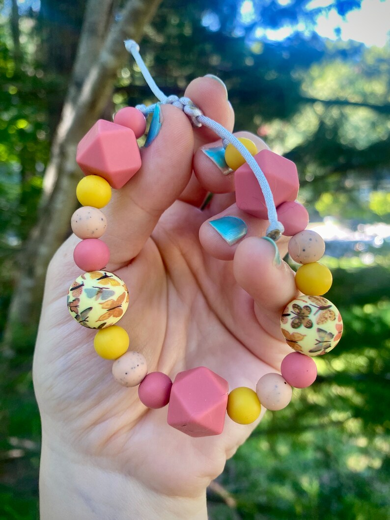 May include: A pink and yellow silicone teething bracelet with a variety of textures and patterns. The bracelet has a knotted cord and is strung with beads that include a geometric shape, a patterned bead, and a solid colour bead.