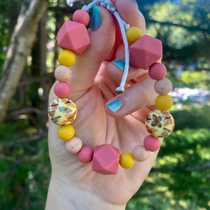 May include: A pink and yellow silicone bracelet with a variety of beads, including some with a leaf pattern.