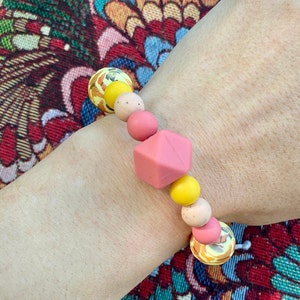 May include: A pink and yellow silicone bracelet with a geometric bead. The bracelet is made of multiple beads, including round, textured, and geometric shapes.