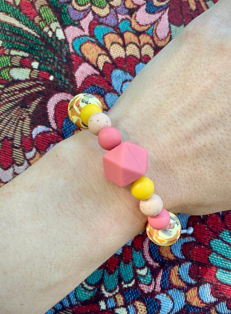 May include: A pink and yellow silicone bracelet with a geometric bead. The bracelet is made of multiple beads, including round, textured, and geometric shapes.