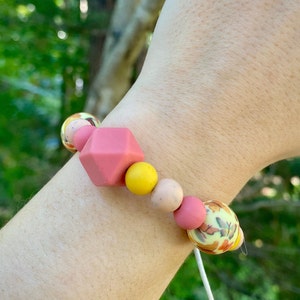 May include: A pink, yellow and beige silicone bracelet with a geometric pink bead and a floral patterned bead. The bracelet is on a person's wrist.