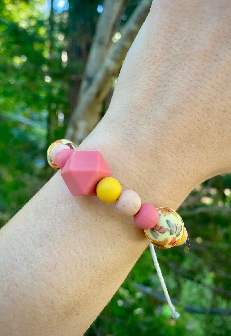 May include: A pink, yellow and beige silicone bracelet with a geometric pink bead and a floral patterned bead. The bracelet is on a person's wrist.