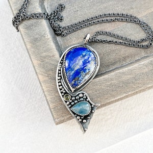 May include: A silver pendant necklace with a blue and green gemstone design. The pendant features a large teardrop-shaped blue gemstone surrounded by smaller green gemstones. The necklace is made of a delicate silver chain.