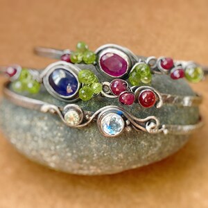 May include: Three silver bracelets with different colored gemstones. The top bracelet has a red gemstone, the middle bracelet has a blue gemstone, and the bottom bracelet has a clear gemstone. All bracelets have green gemstones and are stacked on a gray rock.