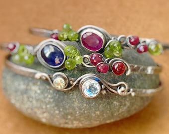 May include: Three silver bracelets with different colored gemstones. The top bracelet has a red gemstone, the middle bracelet has a blue gemstone, and the bottom bracelet has a clear gemstone. All bracelets have green gemstones and are stacked on a gray rock.