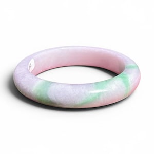 May include: A smooth, round jade bangle bracelet. The bracelet features a blend of colors, including pink, green, and white. The polished surface reflects light, highlighting the natural variations in the stone. This piece is a classic jewelry item.