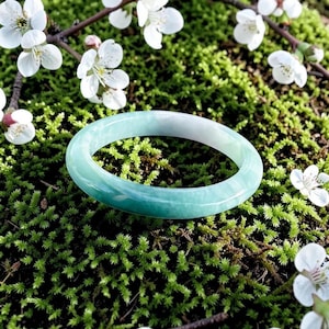 May include: A smooth, polished bangle bracelet featuring a translucent light green and white marbled pattern. The circular band has a rounded profile and a glossy finish, resting against a vibrant green mossy surface with small white flowers.