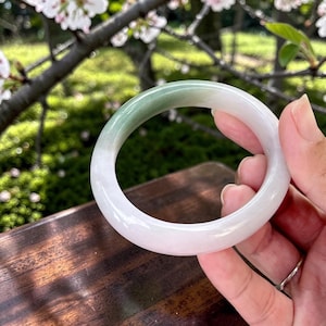 May include: A smooth, circular bangle bracelet crafted from polished stone, featuring a gradient color transition from a translucent, milky white base to a deep, solid forest green section along the upper curve of the rounded band.
