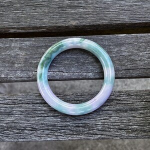 May include: A smooth, circular bangle bracelet crafted from polished stone. The piece features a translucent appearance with a marbled color palette, showcasing blended sections of soft lavender, pale mint green, and deeper emerald green tones.