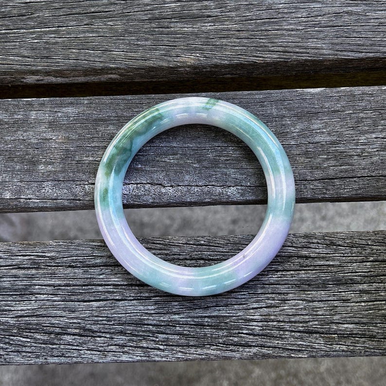May include: A smooth, circular bangle bracelet crafted from polished stone. The piece features a translucent appearance with a marbled color palette, showcasing blended sections of soft lavender, pale mint green, and deeper emerald green tones.