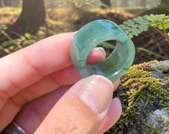 May include: A smooth, polished ring crafted from translucent green jade stone. The band features a wide, rounded profile with natural variations in colour, ranging from light to deep emerald green tones throughout the solid, circular stone structure.