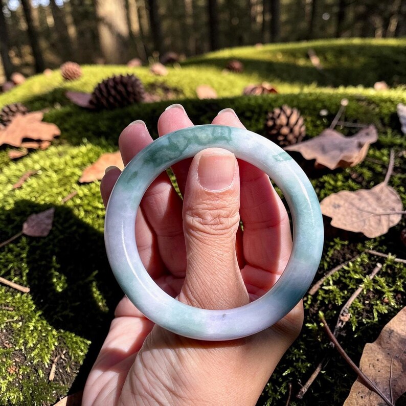 May include: A smooth, circular bangle bracelet featuring a polished surface with a marbled appearance. The stone displays a blend of translucent light green and soft lavender hues, with darker green veining patterns throughout the solid material.
