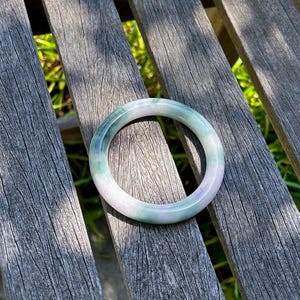 May include: A smooth, circular bangle bracelet crafted from polished stone, featuring a gradient of translucent pale lavender and soft green hues. The solid, continuous band has a rounded profile and a glossy finish that reflects light.