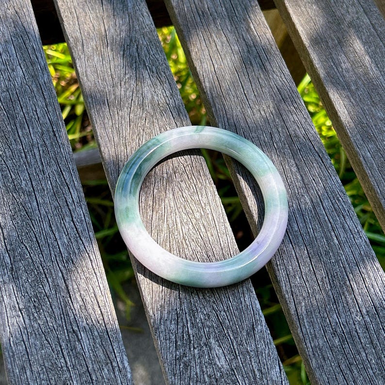 May include: A smooth, circular bangle bracelet crafted from polished stone, featuring a gradient of translucent pale lavender and soft green hues. The solid, continuous band has a rounded profile and a glossy finish that reflects light.