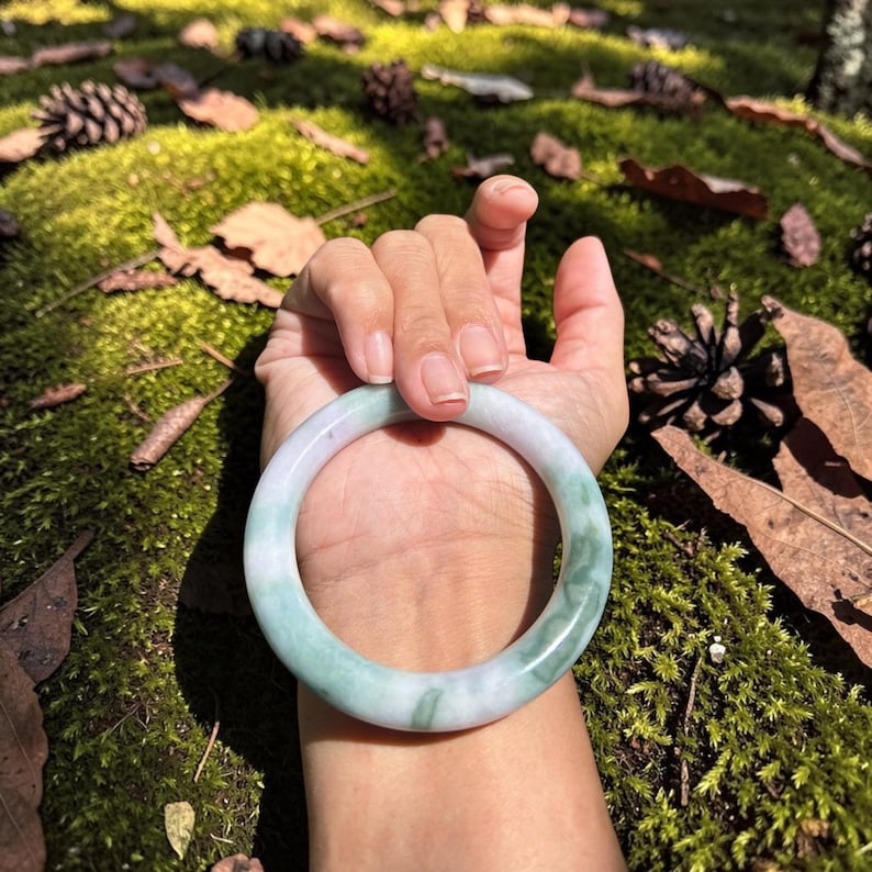 May include: A smooth, circular bangle bracelet crafted from polished stone, featuring a translucent white base with marbled swirls of soft green and lavender throughout the solid, continuous band design.