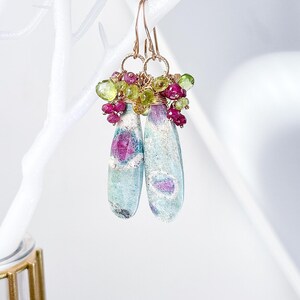 May include: A pair of teardrop-shaped earrings with a blue and green stone set in gold wire with pink and green gemstones surrounding the stone.