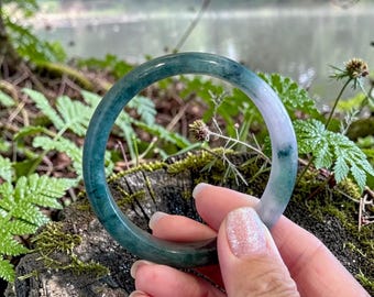 May include: A smooth, circular bangle bracelet crafted from polished stone, featuring a gradient of deep forest green transitioning into a translucent, pale lavender-white hue with natural mineral inclusions throughout the solid material.