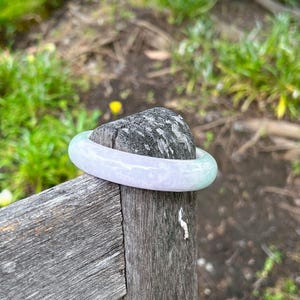 May include: A smooth, polished bangle bracelet crafted from translucent stone, featuring a soft gradient of pale lavender and light green hues. The circular accessory has a rounded profile and a glossy finish that reflects light across its surface.