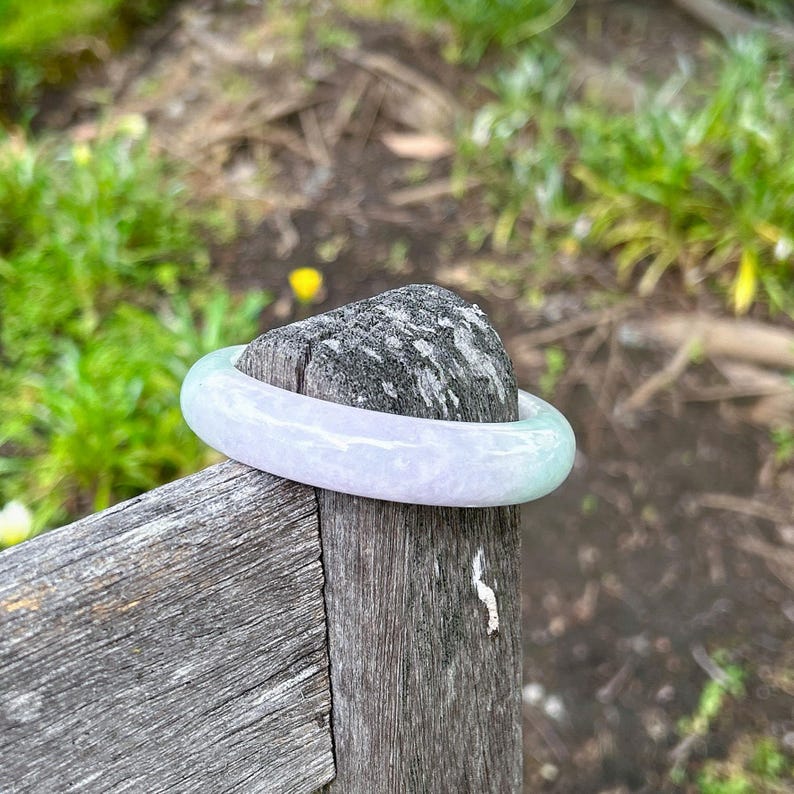 May include: A smooth, polished bangle bracelet crafted from translucent stone, featuring a soft gradient of pale lavender and light green hues. The circular accessory has a rounded profile and a glossy finish that reflects light across its surface.
