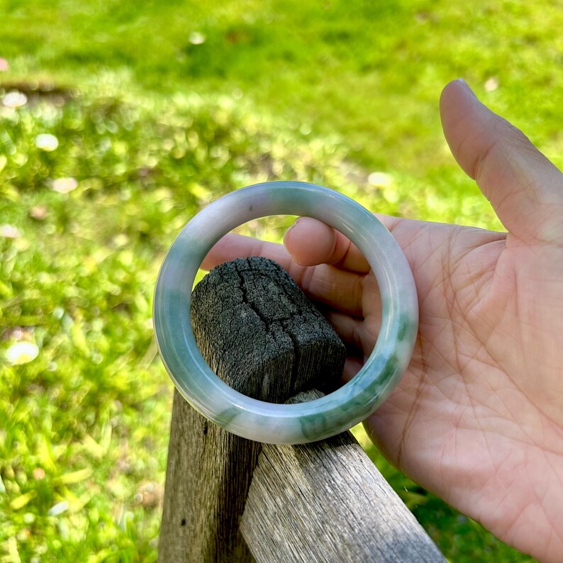 May include: A smooth, circular bangle bracelet featuring a translucent appearance with blended white and green color variations throughout the polished stone surface, showcasing natural mineral patterns and a rounded, continuous band design.