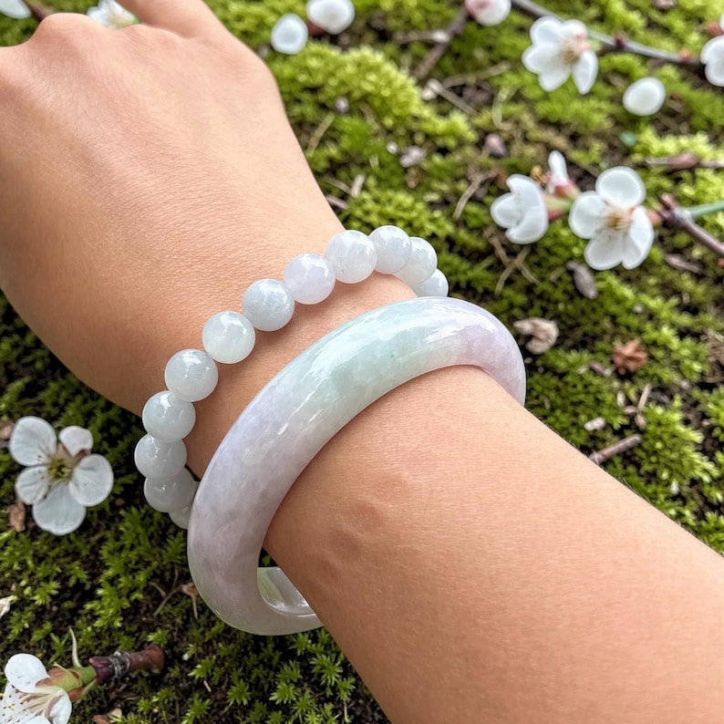 May include: A smooth, solid light green and white jade bangle bracelet paired with a matching beaded jade bracelet featuring round, polished beads. Both pieces exhibit a translucent, marbled appearance with subtle variations in colour and texture.