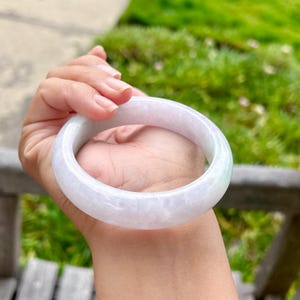 May include: A smooth, polished bangle bracelet crafted from translucent, pale lavender jadeite. The circular accessory features a solid, continuous band with a glossy finish, showcasing natural cloudy white and light purple mineral inclusions throughout.