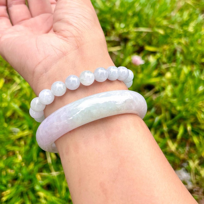 May include: A smooth, solid lavender and pale green jade bangle bracelet paired with a matching beaded jade bracelet featuring round, polished beads. Both pieces exhibit a translucent, light-toned appearance with subtle colour variations throughout.
