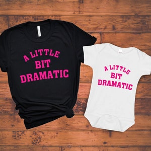 May include: A black t-shirt and a white baby onesie with the text "A Little Bit Dramatic" in pink lettering.