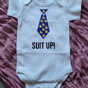 May include: White baby onesie with a blue tie made of yellow rubber ducks. The text "SUIT UP!" is printed below the tie.
