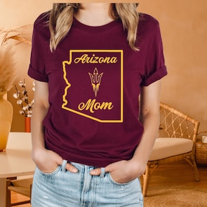 May include: Burgundy t-shirt with a gold outline of the state of Arizona and the words "Arizona Mom" with a gold trident graphic inside the state outline.