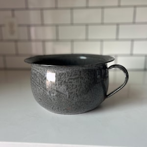 May include: A gray enamelware chamber pot with a handle. The pot has a speckled pattern and a wide rim. The handle is a simple, curved design. The pot is sitting on a white surface.