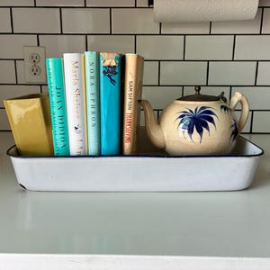 May include: A white rectangular enamel tray with a blue rim containing a yellow ceramic vase, five books with various colored spines, and a cream-colored teapot with a blue palm leaf pattern and a metal lid, all arranged on a white surface.