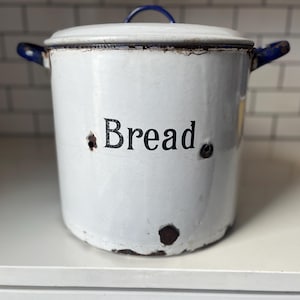May include: White enamel bread bin with a lid and blue accents. The word "Bread" is printed on the front in black. The bin has two blue handles and a blue handle on the lid. The bin has a distressed look.