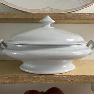May include: A white, lidded serving dish with a rounded base and two handles. The lid has a decorative finial. The dish is made of ceramic or porcelain and is likely vintage. The dish is sitting on a wooden shelf.