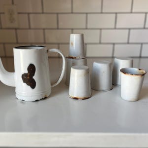 Peut inclure: Théière en émail blanc avec bec et anse, à côté de plusieurs petites tasses en émail blanc. La théière présente un motif floral brun foncé et des signes d'usure. Les tasses sont cylindriques et montrent des signes d'âge.