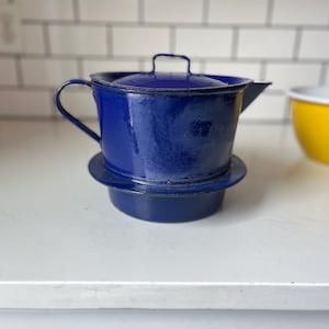 Pode incluir: Um filtro de café esmaltado azul cobalto com tampa e base a condizer. A cafeteira tem uma pega e um bico. Uma tigela amarela está no fundo. Utensílios de cozinha.