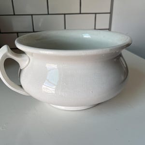 May include: A white ceramic chamber pot with a handle. The pot has a rounded body and a wide rim. The handle is curved and attached to the side of the pot. The pot is sitting on a white surface.