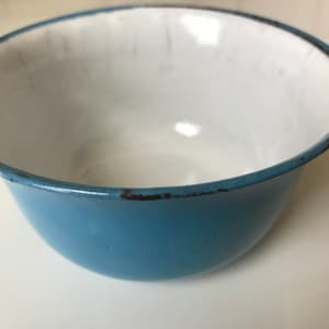 May include: A light blue enamel bowl with a white interior. The bowl has a worn rim and a faded inscription on the bottom.
