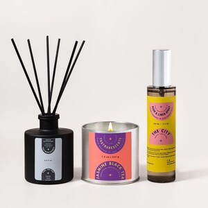 May include: A black reed diffuser, a lit candle, and a room and linen spray. The candle is in a silver tin with a pink and purple label that reads "Jasmine Black Tea." The spray bottle has a yellow label that reads "The City."