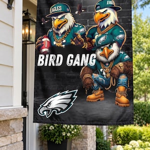 May include: A rectangular garden flag featuring a black brick background with three anthropomorphic eagles wearing teal jerseys, caps, and gold chains. The text BIRD GANG is printed in white, accompanied by an Eagles logo and a football.