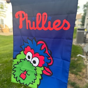 May include: A Phillies team flag featuring the team's name in red script with blue stars, and a cartoon mascot. The flag has a blue background with a green, red, and blue mascot design. The flag is hanging outside.