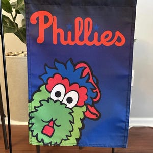 Op de afbeelding: Een blauwe en zwarte tuin vlag met het woord "Phillies" in rode letters en een cartoon mascotte met groene vacht, rode wangen en grote ogen.