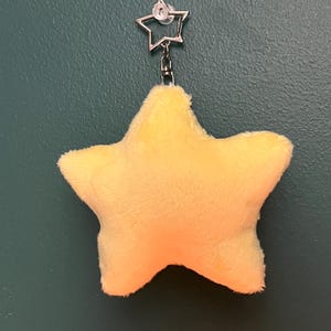 May include: A plush, yellow star-shaped keychain with a silver-colored star-shaped clasp. The star is soft and fluffy, with a gradient of orange at the bottom. The keychain is hanging against a dark teal background.