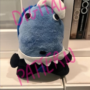 May include: A plush toy in the shape of a blue and white creature with black accents. The toy has a white collar and a white hat. The words "DIGITAL PATTERN" are written across the image in pink.