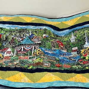 Pode incluir: Um edredom colorido com uma cena de paisagem detalhada com edifícios, uma placa de marina e as palavras "Farmers Market". O edredom tem uma borda azul com padrões geométricos amarelos e verdes. A imagem central retrata uma paisagem urbana vibrante.