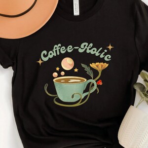May include: Black t-shirt with a graphic of a coffee cup with a green and blue design and the text "Coffee-holic" in a retro font.