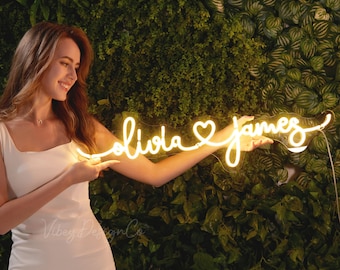 May include: A woman holds a custom neon sign with the names "Olivia" and "James" connected by a heart. The sign is lit up with a warm yellow glow.