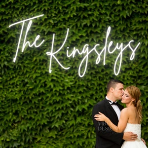 Può includere: Un'insegna al neon bianco brillante con il testo The Kingsleys in un carattere corsivo. L'insegna è montata contro una fitta parete di foglie di edera verde. Le lettere sono illuminate da una luce nitida che risalta sul fogliame.